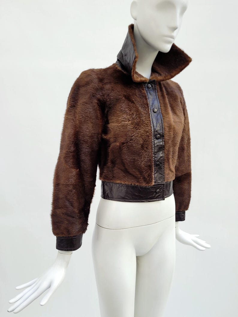 May include: A brown fur jacket with a high collar and leather detailing. The jacket has a dark brown leather collar, button-up front, and cuffs. The waistline and cuffs are trimmed with dark brown leather. The jacket is displayed on a mannequin.