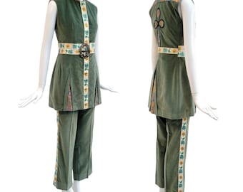 May include: A vintage green velvet two-piece outfit. The sleeveless top and cropped pants are accented with floral trim and a decorative belt. The outfit features a high collar and a unique design on the back of the top. The outfit is displayed on a mannequin.