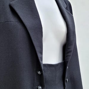 May include: A solid black blazer featuring a notched lapel collar and a front closure with two small black buttons. The garment is displayed on a white mannequin, showcasing the structured fabric texture and the clean lines of the tailored design.