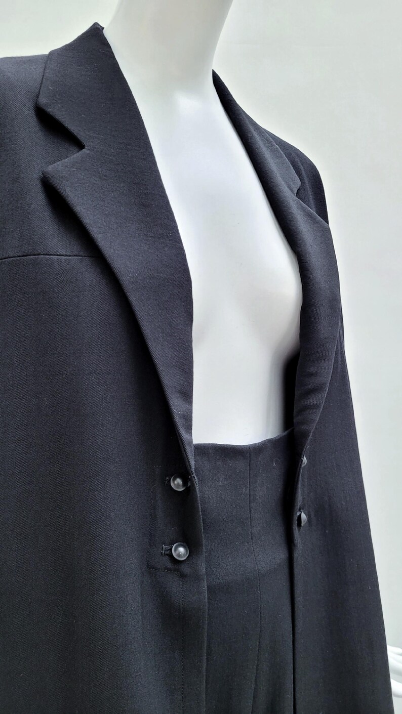 May include: A solid black blazer featuring a notched lapel collar and a front closure with two small black buttons. The garment is displayed on a white mannequin, showcasing the structured fabric texture and the clean lines of the tailored design.