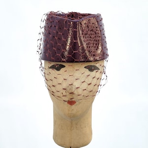 May include: A cylindrical pillbox hat in a deep metallic burgundy with a textured, reptilian-like finish. The hat is adorned with a matching burgundy netting veil that drapes over the front, creating a structured and classic headpiece design.