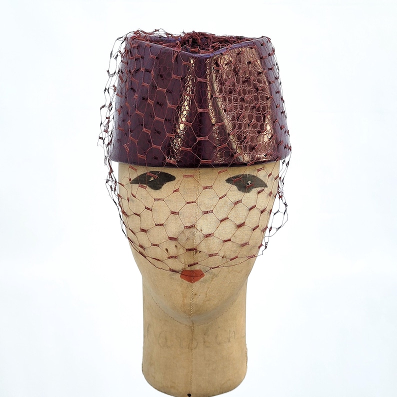 May include: A cylindrical pillbox hat in a deep metallic burgundy with a textured, reptilian-like finish. The hat is adorned with a matching burgundy netting veil that drapes over the front, creating a structured and classic headpiece design.