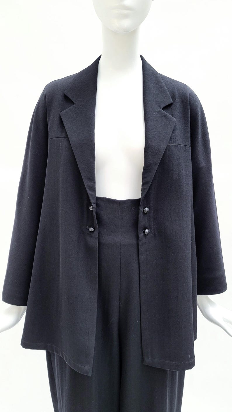 May include: A black blazer featuring a notched lapel collar, long sleeves, and a front closure with two pairs of small black buttons. The jacket has a straight hemline and a structured fit, displayed on a white mannequin against a plain white background.