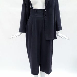 May include: A dark navy blue two-piece set consisting of a long-sleeved blazer and high-waisted cropped trousers. The trousers feature a wide waistband with four small buttons and suspender straps that extend upward from the front waistline.