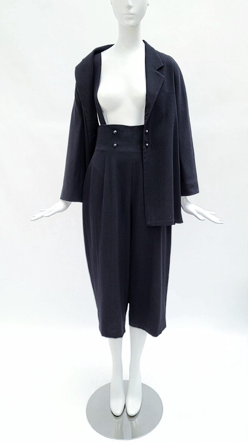 May include: A dark navy blue two-piece set consisting of a long-sleeved blazer and high-waisted cropped trousers. The trousers feature a wide waistband with four small buttons and suspender straps that extend upward from the front waistline.