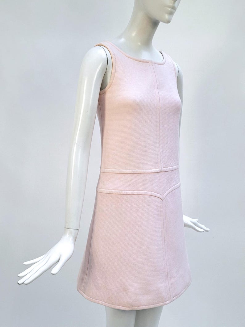 Puede incluir: Un vestido rosa claro sin mangas con cuello redondo y corpi&ntilde;o entallado. El vestido presenta un sutil patr&oacute;n texturizado y una falda acampanada. El vestido se exhibe en un maniqu&iacute;. El vestido es de estilo vintage.