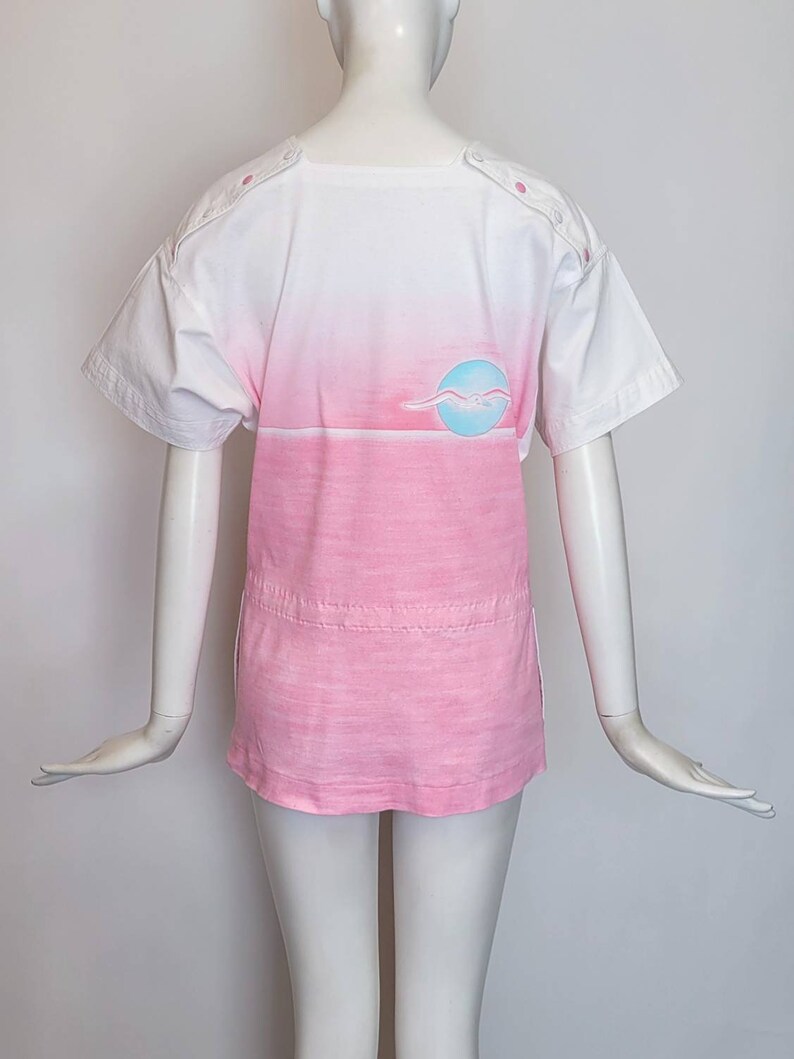 May include: A white and pink ombre t-shirt with a blue and white seagull graphic on the back. The shirt has a round neckline and short sleeves.