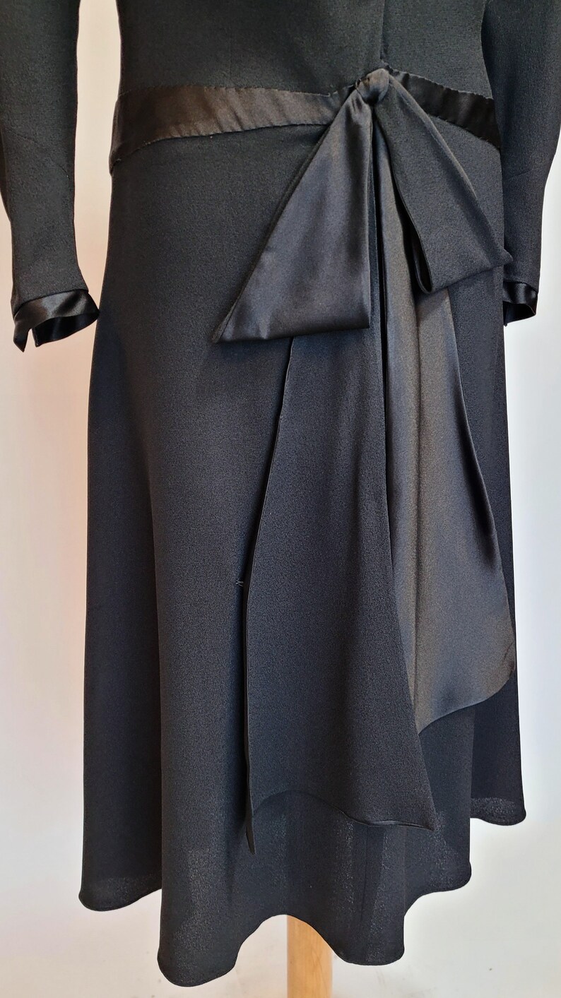 Puede incluir: Un vestido negro con un lazo de sat&eacute;n en la cintura. El vestido tiene un corpi&ntilde;o ajustado y una falda acampanada.