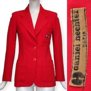 Può includere: Un blazer rosso con una chiusura a un solo bottone e due tasche. Il blazer presenta un piccolo disegno ricamato sulla tasca sinistra. L'etichetta sul blazer recita "Daniel Hechter Paris".