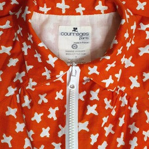 May include: Orange shirt with a white zipper and a white pattern of crosses. The shirt has a label that reads "courreges paris made in france 00 FABRIQUE EN FRANCE MODELE/STYLE 115301".