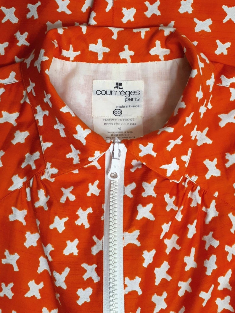 May include: Orange shirt with a white zipper and a white pattern of crosses. The shirt has a label that reads "courreges paris made in france 00 FABRIQUE EN FRANCE MODELE/STYLE 115301".