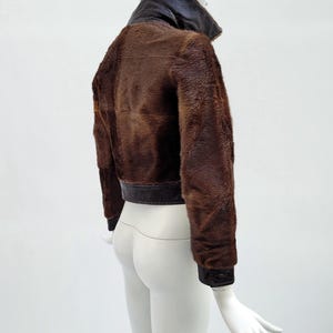 May include: A brown fur jacket with a dark brown leather collar and trim. The jacket has long sleeves and a cropped length. The mannequin is white, and the background is plain white.
