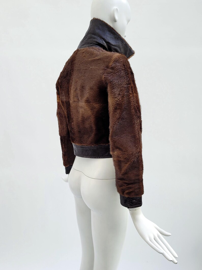May include: A brown fur jacket with a dark brown leather collar and trim. The jacket has long sleeves and a cropped length. The mannequin is white, and the background is plain white.