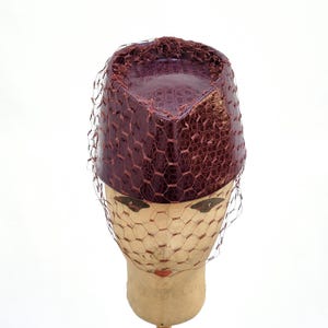 May include: A cylindrical pillbox hat in a deep burgundy colour with a textured, faux-reptile skin pattern. The hat is adorned with a fine, dark brown netting veil that drapes over the sides and front, creating a structured and layered appearance.