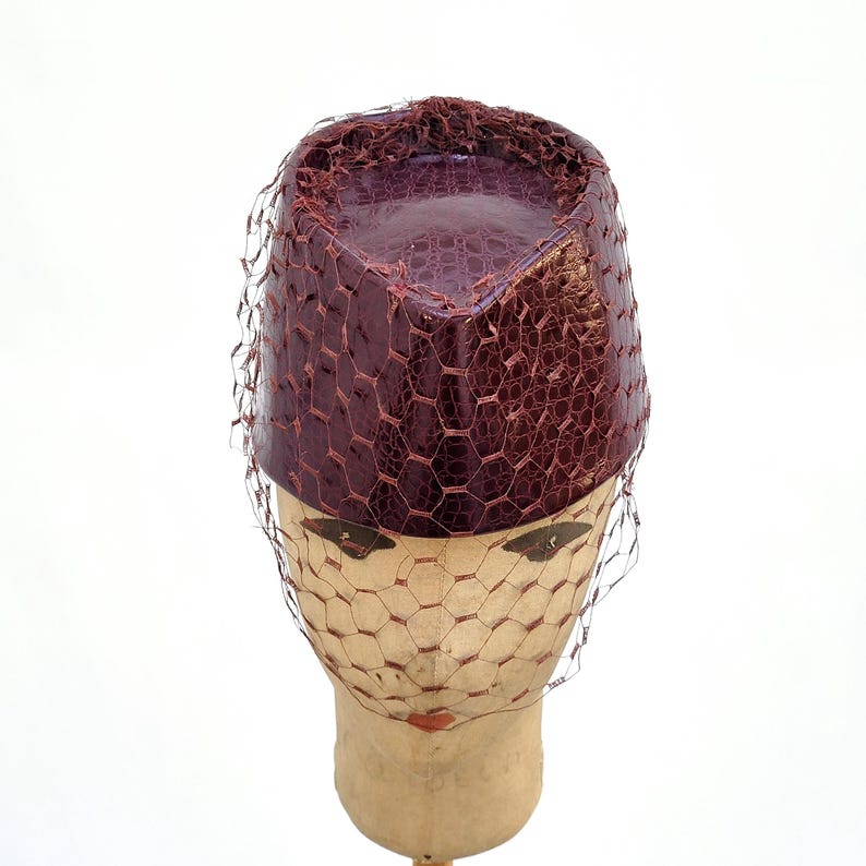 May include: A cylindrical pillbox hat in a deep burgundy colour with a textured, faux-reptile skin pattern. The hat is adorned with a fine, dark brown netting veil that drapes over the sides and front, creating a structured and layered appearance.