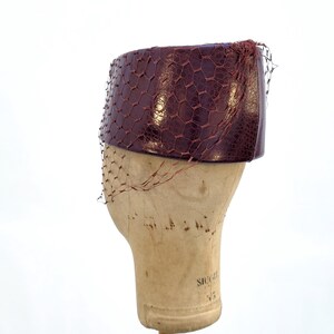 May include: A deep burgundy pillbox hat with a textured, reptilian-like surface pattern. The hat is adorned with a fine, matching burgundy netting that drapes over the sides, creating a hexagonal mesh overlay across the structured, cylindrical form.