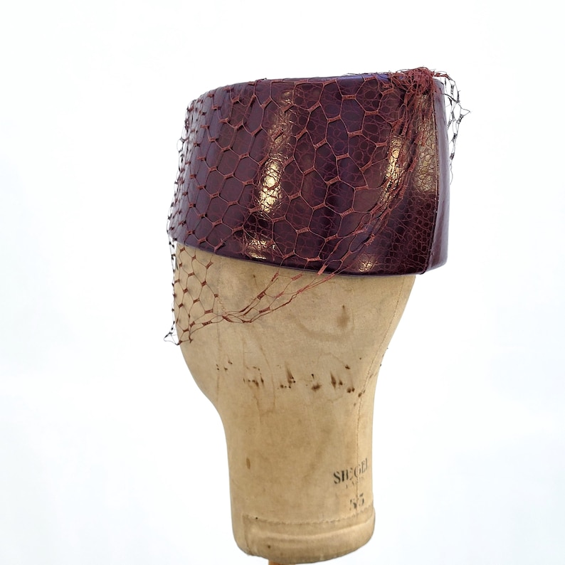 May include: A deep burgundy pillbox hat with a textured, reptilian-like surface pattern. The hat is adorned with a fine, matching burgundy netting that drapes over the sides, creating a hexagonal mesh overlay across the structured, cylindrical form.