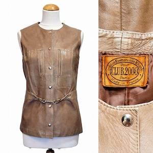 Peut inclure: Un gilet en cuir marron avec un devant à boutons et une ceinture avec une boucle en métal. Le gilet porte une étiquette qui indique "Création Emmanuelle Khann Pour Club 2000 Paris Fabriqué en France".
