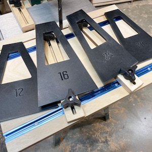 May include: Four black wooden jigs with different sized openings, labeled 12, 16, 20, and 24, are attached to a wooden table with blue tracks. The jigs are used for woodworking projects.