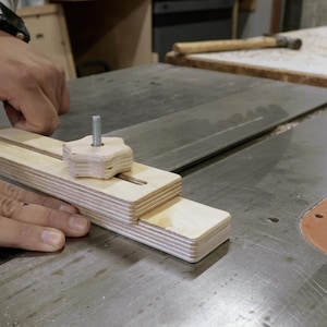 May include: A wooden jig with a star-shaped knob and a metal bolt is positioned on a table saw. The jig is made of light-colored wood and is designed to assist with woodworking tasks. The saw blade is visible.