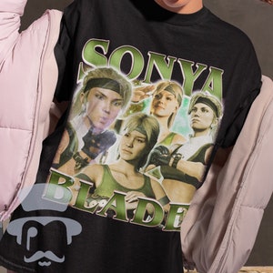 May include: Black cotton t-shirt with a graphic of Sonya Blade from Mortal Kombat. The design features multiple images of the character with the text "SONYA BLADE" in a green, vintage-style font. The shirt has the text "METALCORPSZ Cotton Shirt" at the bottom.