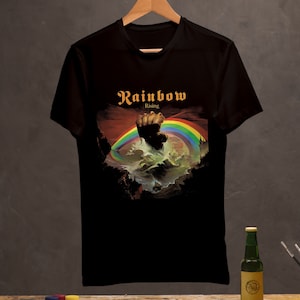 Puede incluir: Camiseta de algodón negra con un gráfico de un arcoíris y un puño, con el texto "Rainbow Rising" en la parte delantera.