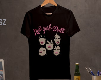May include: Black cotton t-shirt with a pink graphic of the New York Dolls band name and five faces. The band name is written in a cursive font.