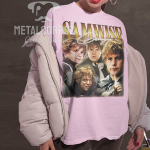 May include: A pink t-shirt with a vintage-style graphic of Samwise Gamgee from the Lord of the Rings movies. The graphic features four images of Samwise Gamgee in different poses. The text "Samwise Gamgee" is printed in gold letters above the graphic.