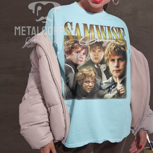 May include: A light blue t-shirt with a vintage-style graphic of Samwise Gamgee from the Lord of the Rings. The graphic features four images of Samwise Gamgee in different poses. The text "SAMWISE Gamgee" is printed in gold letters above the graphic.