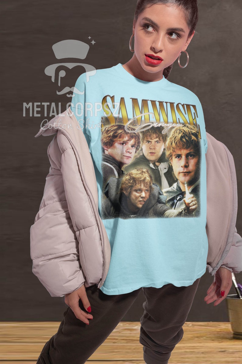May include: A light blue t-shirt with a vintage-style graphic of Samwise Gamgee from the Lord of the Rings. The graphic features four images of Samwise Gamgee in different poses. The text "SAMWISE Gamgee" is printed in gold letters above the graphic.