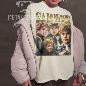 May include: A white t-shirt with a vintage-style graphic of Samwise Gamgee from the Lord of the Rings. The graphic features four images of Samwise Gamgee in different poses. The text "SAMWISE Gamgee" is printed in gold letters above the graphic.