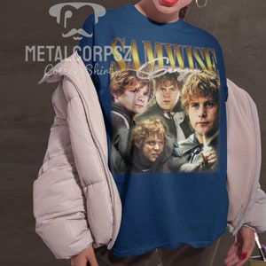 May include: A navy blue t-shirt with a vintage-style graphic of Samwise Gamgee from the Lord of the Rings. The graphic features four images of Samwise Gamgee in different poses. The text "SAMWISE GAMGEE" is printed in gold letters above the graphic.