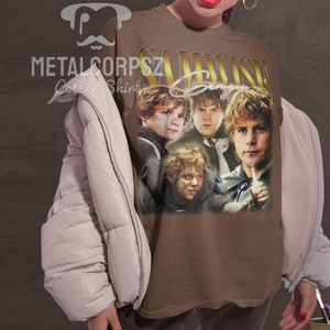 May include: A brown t-shirt with a vintage-style graphic of Samwise Gamgee from the Lord of the Rings. The graphic features four images of Samwise Gamgee in different poses. The text "Samwise Gamgee" is printed in gold letters above the graphic.