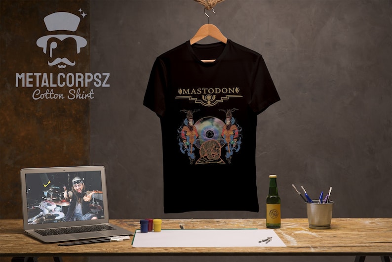 May include: Black cotton t-shirt with a gold graphic of the band Mastodon's logo and album art. The logo reads "Mastodon" in a gold font.