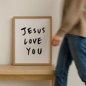 May include: A framed print with the words "JESUS LOVE YOU" in black brush lettering. The print is in a light wood frame and rests on a light wood table. A person in a brown jacket and blue jeans is blurred in the background.