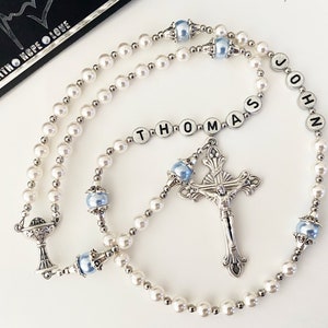 May include: A rosary featuring white pearl beads, light blue accent beads, and silver-toned metal spacers. The rosary includes a silver crucifix and a chalice charm. Letter beads spell out the name THOMAS JOHN in the centre of the strand.