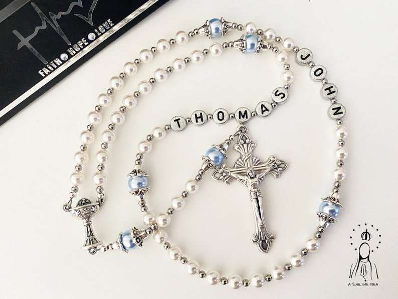 May include: A rosary featuring white pearl beads, light blue accent beads, and silver-toned metal spacers. The rosary includes a silver crucifix and a chalice charm. Letter beads spell out the name THOMAS JOHN in the centre of the strand.
