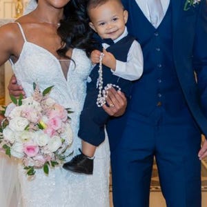 May include: A wedding photograph of a smiling couple and a child. The bride wears a white, embellished gown and holds a bouquet. The groom is in a navy suit, and the child wears a navy suit and holds a rosary.