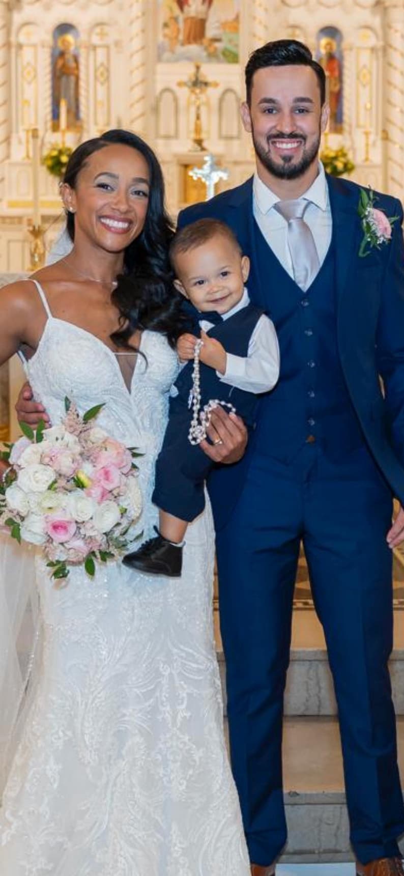 May include: A wedding photograph of a smiling couple and a child. The bride wears a white, embellished gown and holds a bouquet. The groom is in a navy suit, and the child wears a navy suit and holds a rosary.
