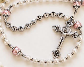 May include: A rosary featuring white pearl-like beads, pink accent beads, and silver-toned metal spacers. The beads spell out the name CHARLOTTE in black letters on silver-toned circular beads. A silver-toned crucifix with an INRI inscription is attached.