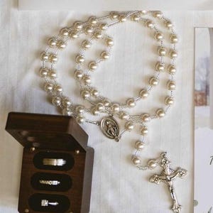 May include: A rosary featuring faux pearl beads and a silver crucifix. A dark wooden ring box contains three rings. The rosary and rings are displayed on a white surface, possibly for a religious ceremony or wedding.
