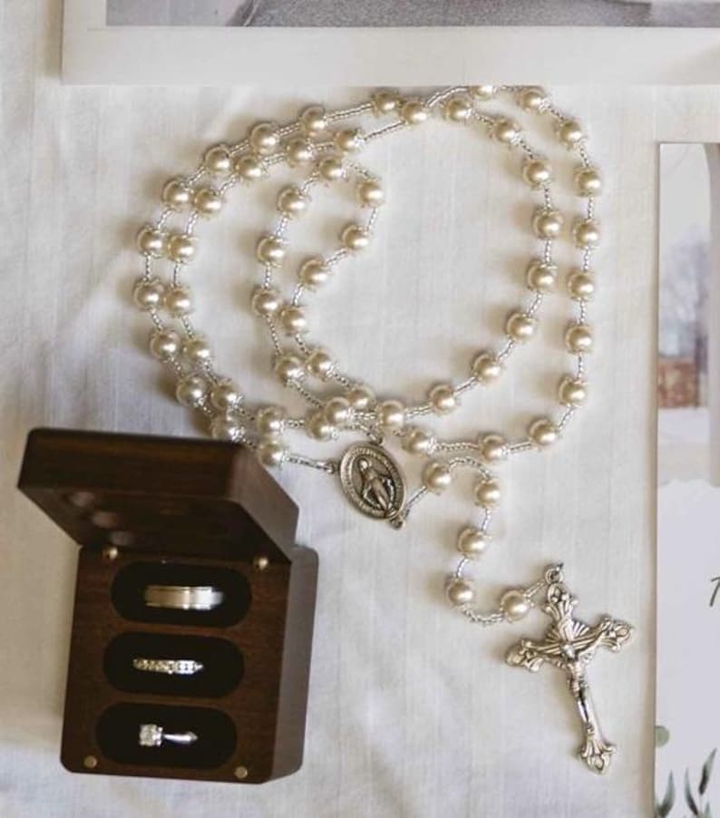 May include: A rosary featuring faux pearl beads and a silver crucifix. A dark wooden ring box contains three rings. The rosary and rings are displayed on a white surface, possibly for a religious ceremony or wedding.