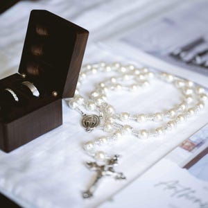 May include: A dark wooden ring box containing three rings, alongside a pearl rosary with a silver crucifix. The rings include a diamond engagement ring and two wedding bands. The rosary rests on a white surface.