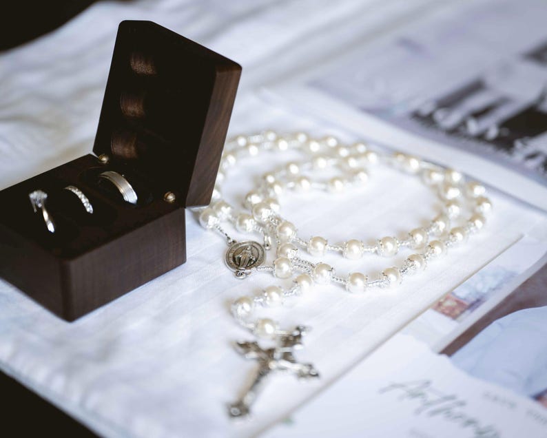 May include: A dark wooden ring box containing three rings, alongside a pearl rosary with a silver crucifix. The rings include a diamond engagement ring and two wedding bands. The rosary rests on a white surface.