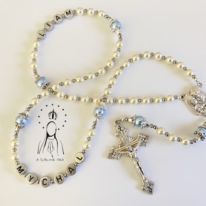 May include: A rosary featuring white pearl-like beads, metallic silver spacer beads, and light blue accent beads. The rosary includes silver letter beads spelling LIAM and MYCHAL, a silver crucifix, and a centrepiece. A logo with text A SUBLIME IDEA is visible.