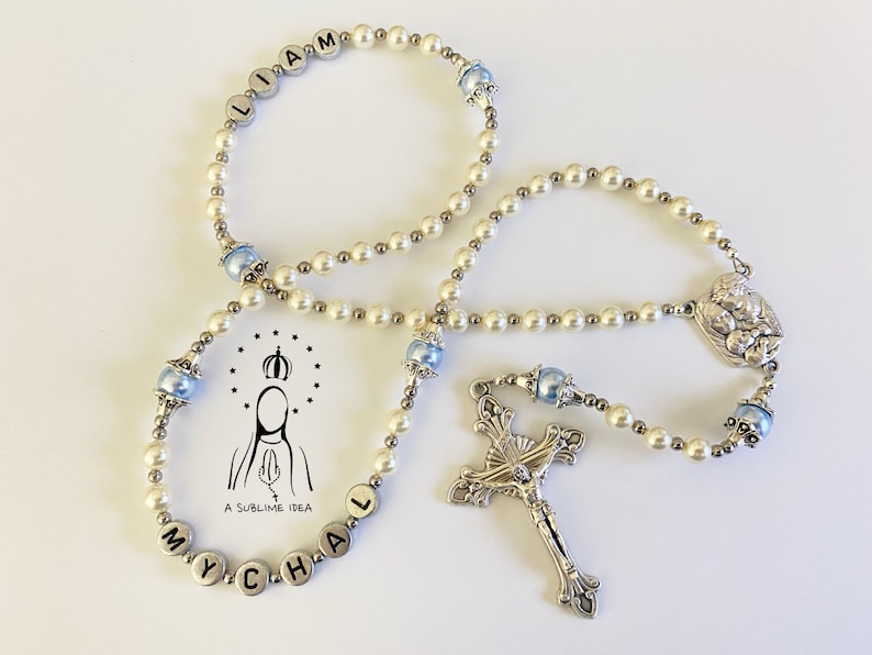 May include: A rosary featuring white pearl-like beads, metallic silver spacer beads, and light blue accent beads. The rosary includes silver letter beads spelling LIAM and MYCHAL, a silver crucifix, and a centrepiece. A logo with text A SUBLIME IDEA is visible.
