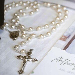 May include: A pearl rosary with a silver crucifix and a medal. The rosary is laid on a white surface next to a wedding invitation. The invitation has the name "Anthony" written on it.