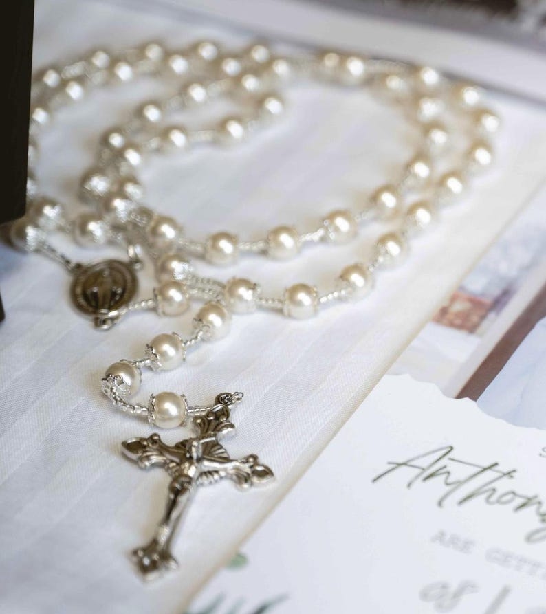 May include: A pearl rosary with a silver crucifix and a medal. The rosary is laid on a white surface next to a wedding invitation. The invitation has the name "Anthony" written on it.