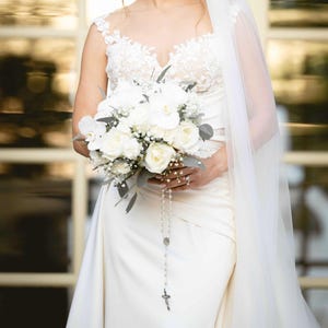 May include: A bride in a white wedding dress holds a bouquet of white roses, orchids, and greenery. A rosary hangs from the bouquet. The dress has a lace bodice and a flowing skirt. A sheer veil drapes over the bride's shoulder.