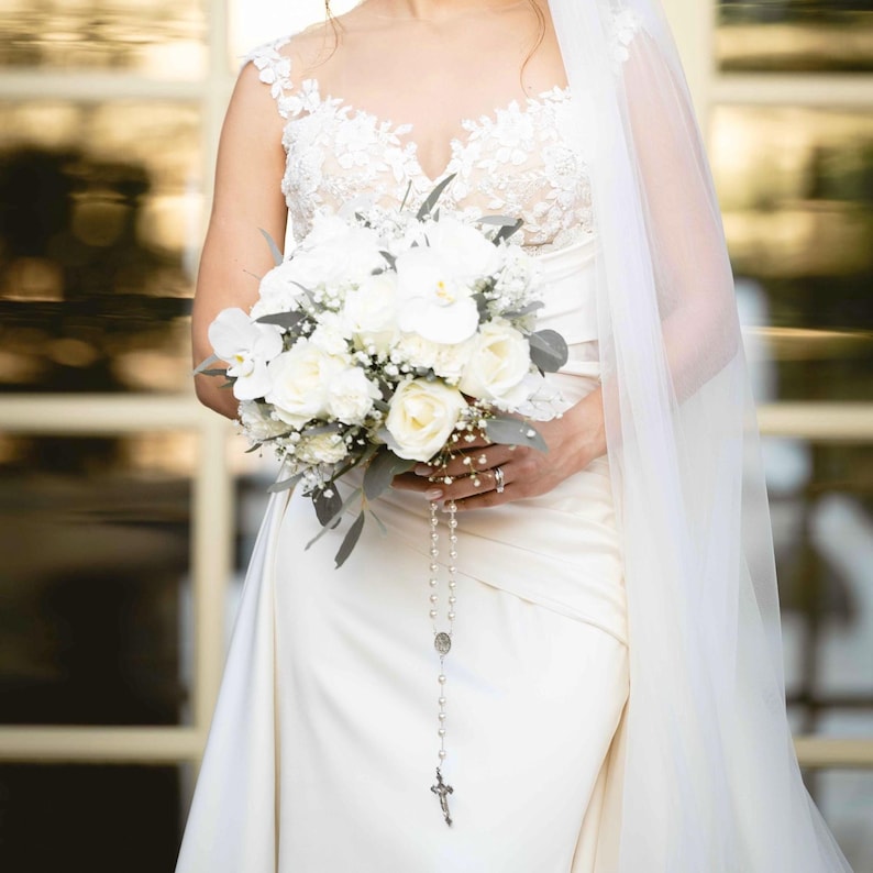 May include: A bride in a white wedding dress holds a bouquet of white roses, orchids, and greenery. A rosary hangs from the bouquet. The dress has a lace bodice and a flowing skirt. A sheer veil drapes over the bride's shoulder.